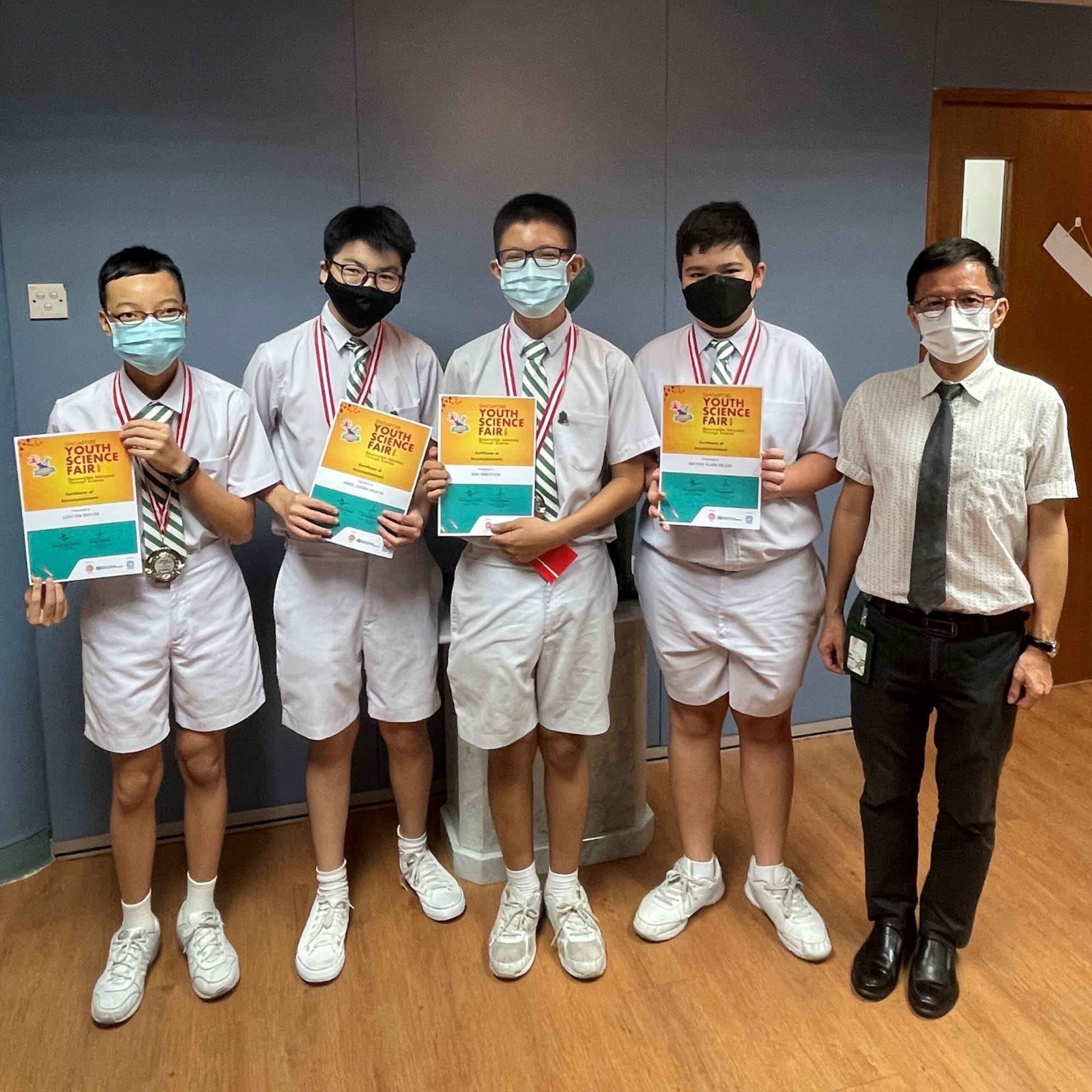 Four masked students holding Youth Science Fair certificates with medals stand beside a masked adult teacher.